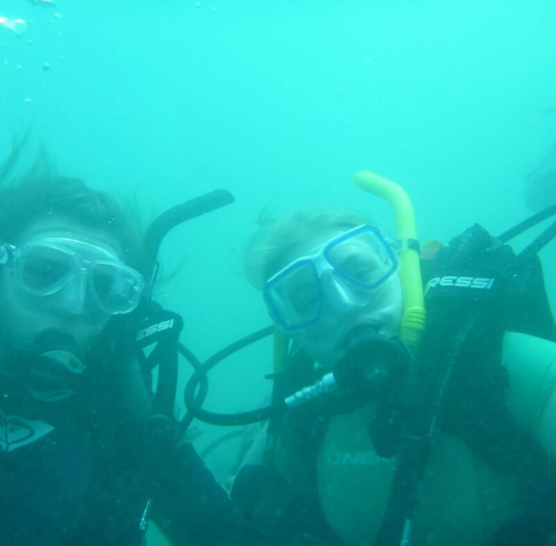 The image shows two people scuba diving. They are both wearing scuba masks and gear, and they appear to be underwater. The water is a murky green color, and there are some bubbles visible in the background. The divers are close to the camera, and their faces are visible. They seem to be enjoying their dive.